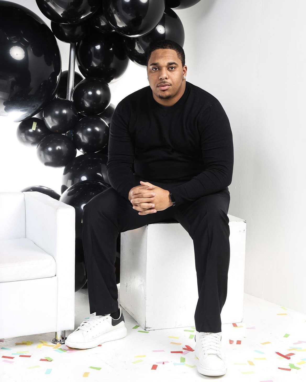Portrait of the photographer dressed in all black, seated against a black balloon backdrop
