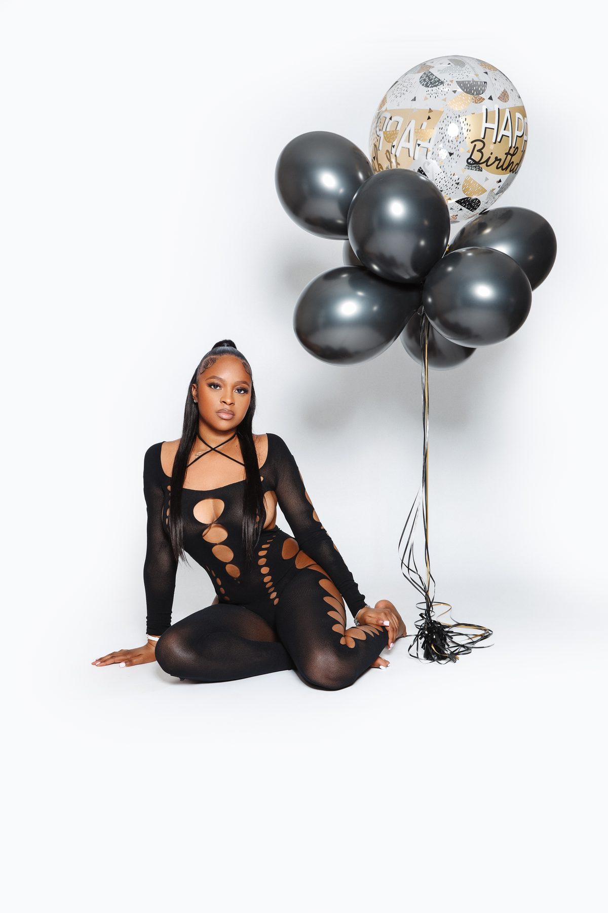 Model in a black cut-out bodysuit seated beside a cluster of birthday balloons on a white set